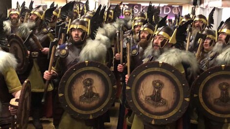 A group of people in Viking-style costumes with helmets, shields, and axes, gathered indoors, looking forward.