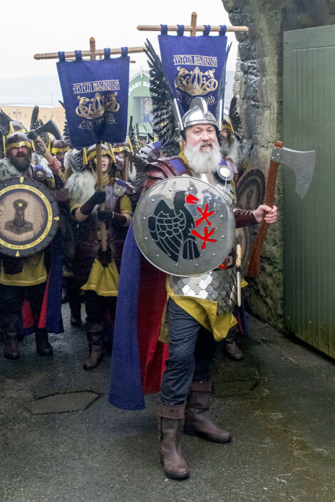 A group of people dressed as Vikings parade with shields, axes, and banners. The leader, in front, wears a helmet and has a beard. They are walking on a paved street.