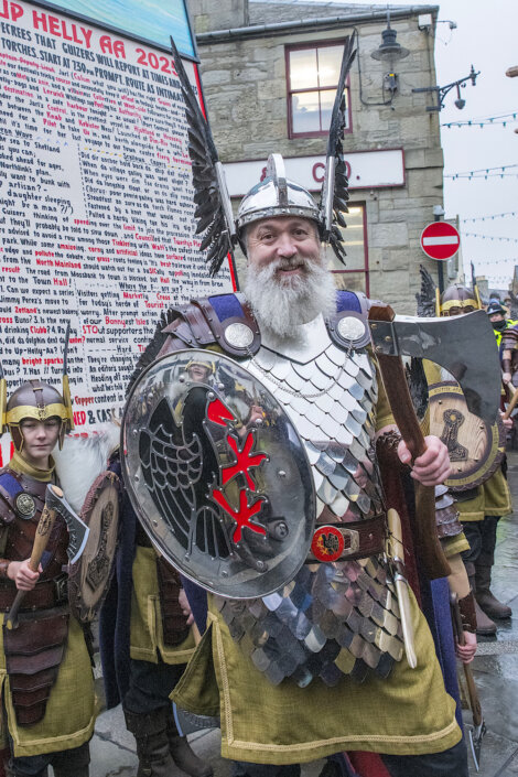 A person in Viking costume with a winged helmet and shield stands outdoors. Behind them are more people in similar attire and a large banner.