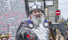 A man in Viking attire with a silver helmet, holding a shield and axe, stands in front of others in similar costumes. A building and a crowd are visible in the background.