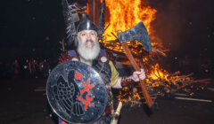 A person dressed in medieval armor with a horned helmet holds a large axe and a shield in front of a large bonfire at night.