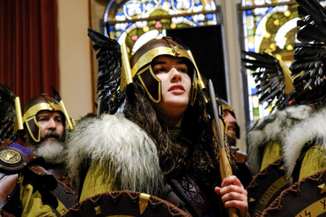 Person in Viking-style costume with helmet and fur accents stands indoors near stained glass windows.