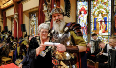 An older woman in ceremonial attire and a man in Roman soldier costume pose with a scroll inside a room with stained glass windows.