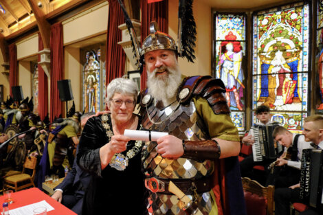 An older woman in ceremonial attire and a man in Roman soldier costume pose with a scroll inside a room with stained glass windows.