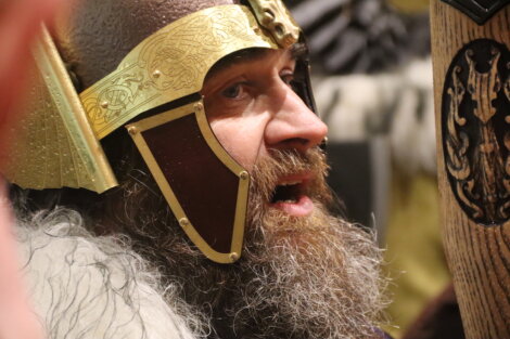 Man dressed in Viking attire with a detailed helmet and a long beard, holding a wooden shield.