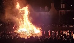 A large bonfire with a wooden structure burning intensely at night, surrounded by a crowd of people watching the event.