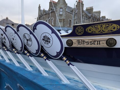 Close-up of a blue and white boat with shields and oars bearing intricate designs, moored near stone buildings.