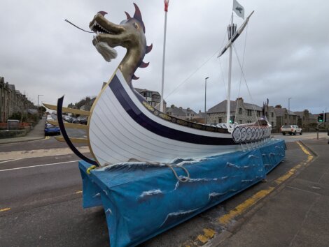 Large dragon-headed Viking ship model on a blue platform, displayed on a street with buildings in the background.