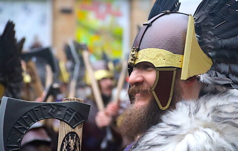 Person in Viking costume with helmet and axe, surrounded by others in similar attire at an outdoor gathering.