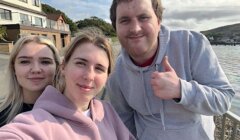 Three people in hoodies pose for a selfie with a waterfront and houses in the background. One person gives a thumbs-up.