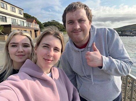 Three people in hoodies pose for a selfie with a waterfront and houses in the background. One person gives a thumbs-up.