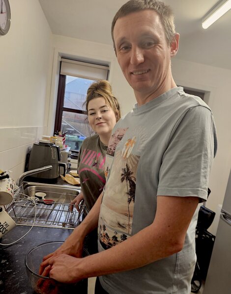 Two people stand in a kitchen. One is mixing something in a bowl while the other looks at the camera. Kitchen appliances are visible in the background.