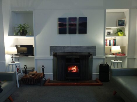 A cozy living room features a lit fireplace, two lamps, a stack of logs, and a plant. Shelves hold books and decor. Paintings hang above the fireplace, and armchairs flank the scene.