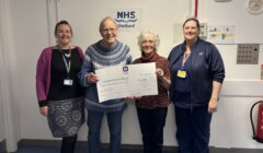 Four people stand indoors, holding a large presentation check for £10,000 addressed to Shetland Health Board. They are smiling and standing in front of an NHS Shetland sign.