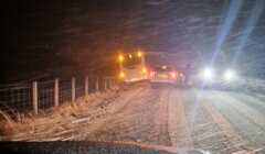 Two cars stopped on a snowy road at night, one slightly off the road near a fence, with headlights and taillights on, surrounded by falling snow.