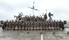 A large group of people dressed as Vikings, holding shields and axes, poses in front of a ship's bow on a rainy day near the sea.