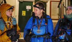 Three people wearing outdoor gear and headlamps talk indoors, preparing for an activity.