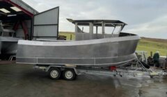 A partially constructed metal boat on a trailer outside a workshop, with overcast skies and grassy hills in the background.