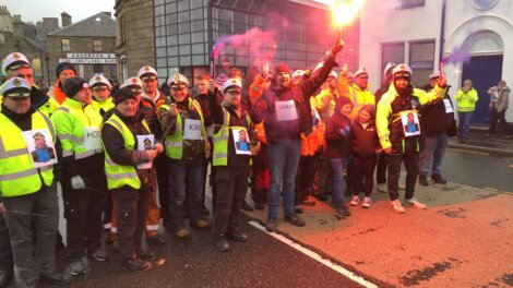 A group of people in high-visibility jackets and sailor hats stand on a street, some holding flares. They hold photos of a person. Buildings in the background.