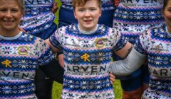 Group of rugby players in patterned uniforms, arms linked, standing on grass. Central player has light brown hair and is wearing a bandage on one arm.