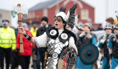 Person in Viking-inspired attire with a shield and axe leading a group in costume during an outdoor event.