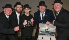 Five men in suits and hats pose with a young person. They are smiling around an open briefcase displaying a painting of a ship in a stormy sea.