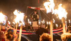 Person dressed as a Viking on a boat float, surrounded by people holding torches during a nighttime event.