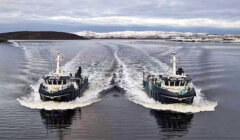 Two boats navigate parallel paths on a calm body of water with snow-covered land in the background, leaving symmetrical wakes behind them.