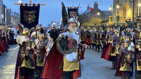 A group of people in Viking costumes march down a street, led by a person holding a shield and wearing a helmet with wings. Banners are visible in the background.