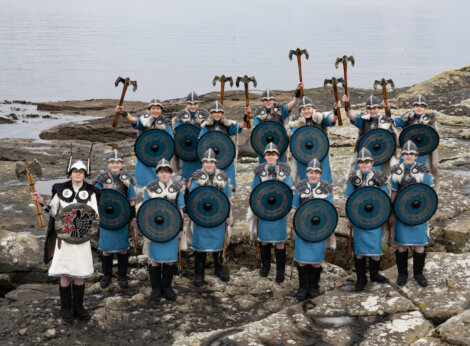 A group of people in Viking costumes stand on rocky terrain near water, holding shields and axes, dressed in blue tunics and helmets.