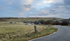 A winding road curves through a rural landscape with grassy fields, a small village in the distance, and partly cloudy skies overhead.