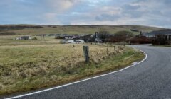 A winding road curves through a rural landscape with grassy fields, a small village in the distance, and partly cloudy skies overhead.