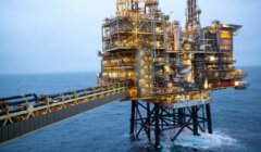 An offshore oil rig stands tall in the ocean, connected by a bridge. The structure is illuminated, highlighting intricate machinery against a backdrop of cloudy skies and calm waters.