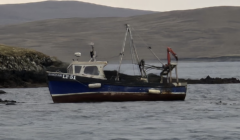 A small blue and white fishing boat is anchored near a rocky shoreline, with hilly terrain in the background.