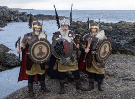 Three people in Viking costumes stand on a rocky shore, each holding a shield and wearing a helmet with horns and feathers. The sea and rocks are visible in the background.