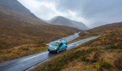 A van drives along a narrow road in a mountainous landscape, with a river running parallel in overcast weather.