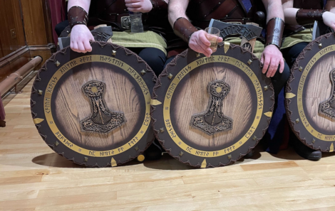 People sitting, each holding a round shield with wooden texture and intricate designs. The shields have Norse symbols and inscriptions around the edge.