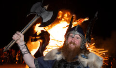 Person in Viking attire holds an axe and smiles in front of a burning structure at night.