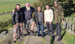 Five men standing on a stone path outdoors, one wearing camo pants, another in a camo jacket, with trees and a field in the background.