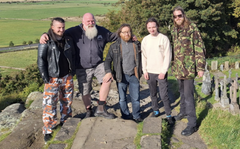 Five men standing on a stone path outdoors, one wearing camo pants, another in a camo jacket, with trees and a field in the background.