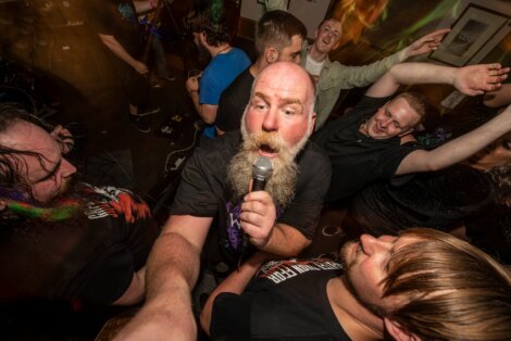 Bearded man with microphone singing energetically among a lively crowd indoors.