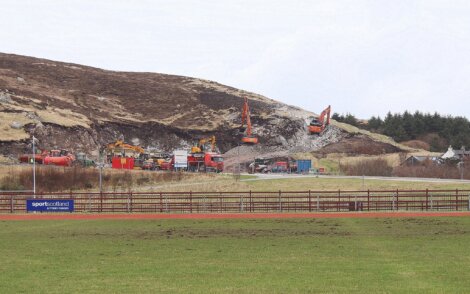 Construction site with several excavators, trucks, and equipment working on a hillside. A grassy field and a small building are visible in the foreground.