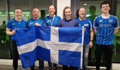 Six people standing indoors, smiling and holding a large blue and white flag with a cross design. They are wearing various blue tops.