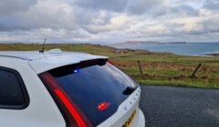 A white Volvo car with rear lights on is parked on a coastal road, overlooking grassy fields and the sea under a cloudy sky.