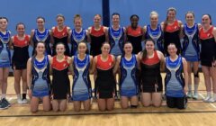 A group of women in team uniforms pose for a photo in a sports hall, with a mix of red and blue designs on their outfits.