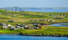 Scenic view of a coastal village with scattered houses, green fields, and surrounding water, with distant cliffs under a clear sky.