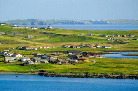 Scenic view of a coastal village with scattered houses, green fields, and surrounding water, with distant cliffs under a clear sky.