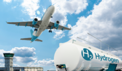 Airplane flying over an airport near a large hydrogen fuel tank marked with "H₂ Hydrogen" under a blue sky with clouds.