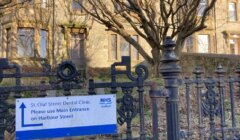 Sign for St. Olaf Street Dental Clinic advises using the main entrance on Harbour Street. Ornate black metal fence and building in the background.