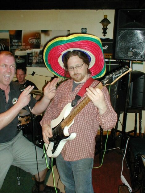 A man wearing a colorful sombrero plays an electric guitar while another man enthusiastically poses beside him. A drummer is visible in the background.